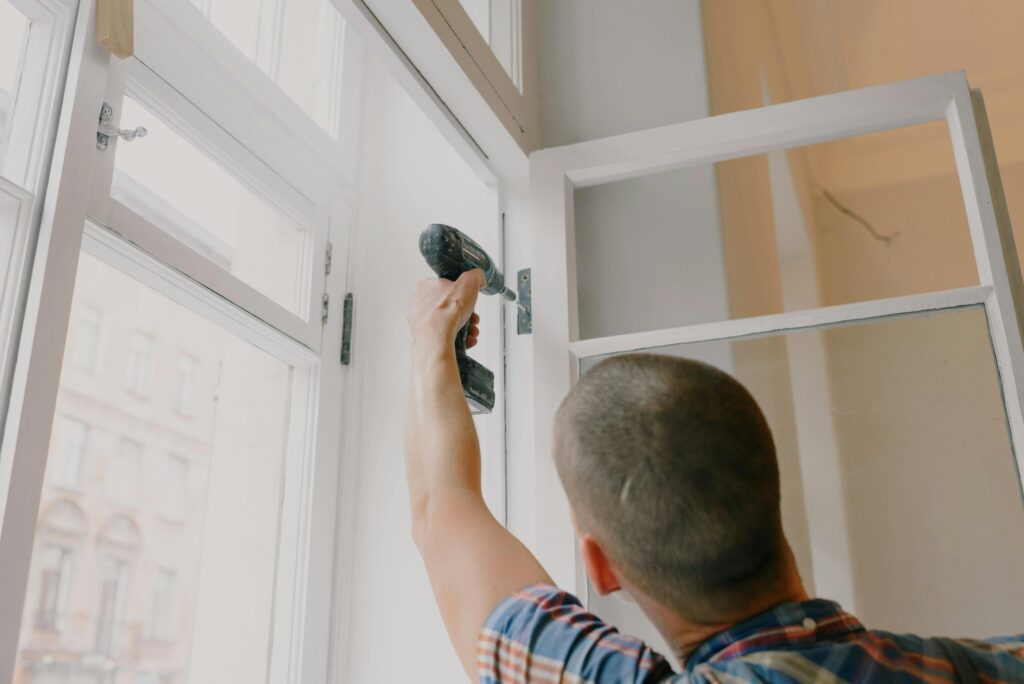 Back view of unrecognizable craftsman using cordless screwdriver and assembling window while screwing metal hinge during renovation works in apartment
